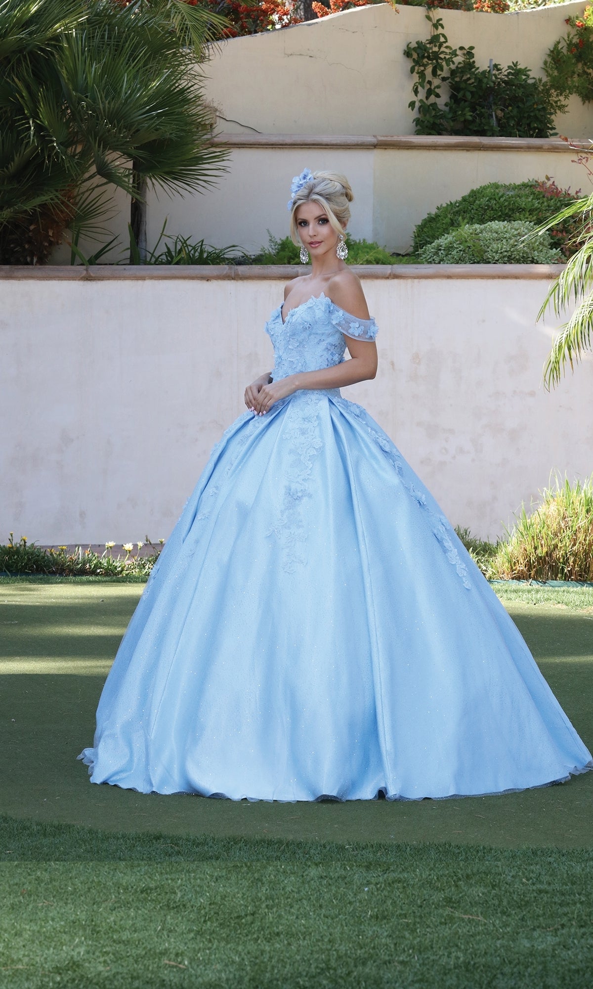 A woman stands on grass wearing the Quinceanera Dress 1501 by Dancing Queen, an off-the-shoulder light blue a-line ball gown with floral details and a matching headpiece. Greenery and a white wall are in the background.